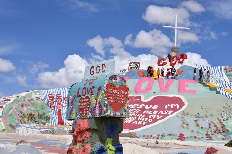 cross, niland, nature, ramp, built structure, slab city, text, sunlight, people, painted mountain, pastel, outdoors, information sign, script, crowd, travel destinations
