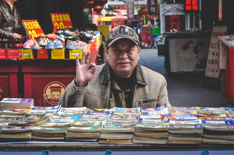 2K, elderly, salesperson, seller, retail, sales, market stall, coat, market, storybooks, casual clothing, salesman, choice, stack, business, indoors, china