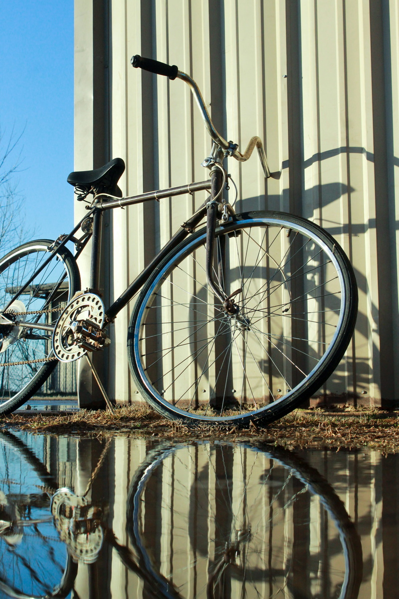 puddle, spoke, tire, outdoors, water, land vehicle, sunlight, wheel, strap, transportation, harsh, shadow, no people, leaning, mountain bike, brake, parking