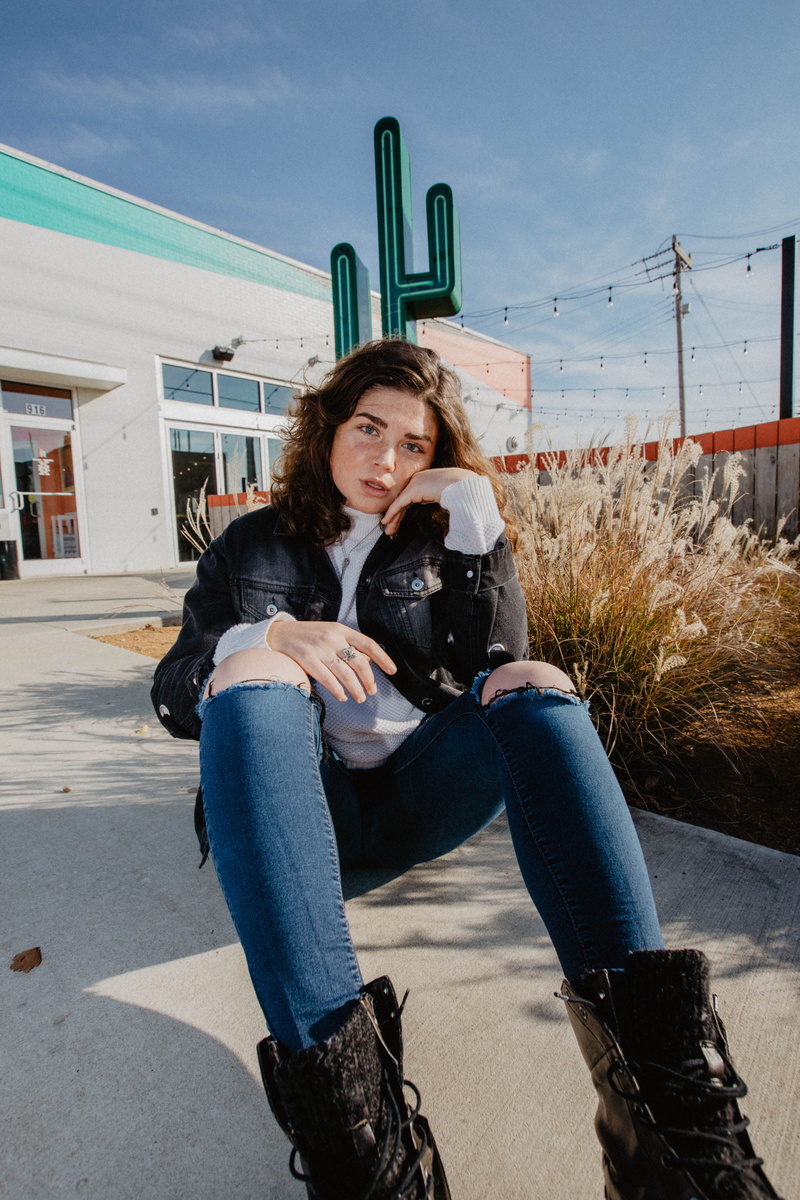 looking at camera, sitting, female, denim, front view, human, store, ground, footwear, shoe, beautiful woman, sky, sign, pose, built structure, 5K, apparel