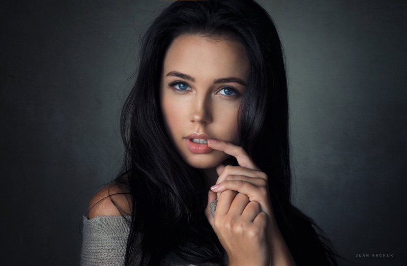young adult, hairstyle, close-up, finger on lips, simple background, hair, looking at camera, headshot, fashion, blue eyes, beauty, black background, hand