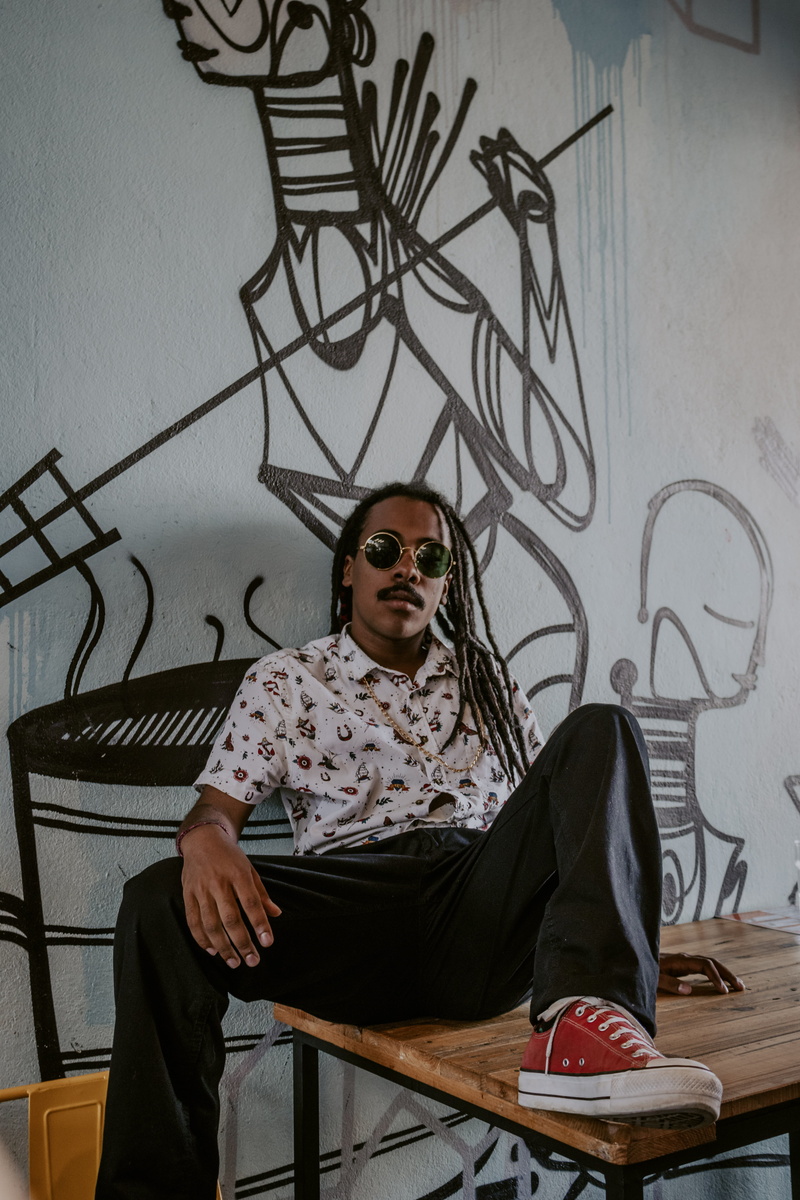 male, fashion, wall, sit, wear, facial expression, leisure activity, room, sunglasses, front view, pose, indoors, black, shirt, model, real people, painting
