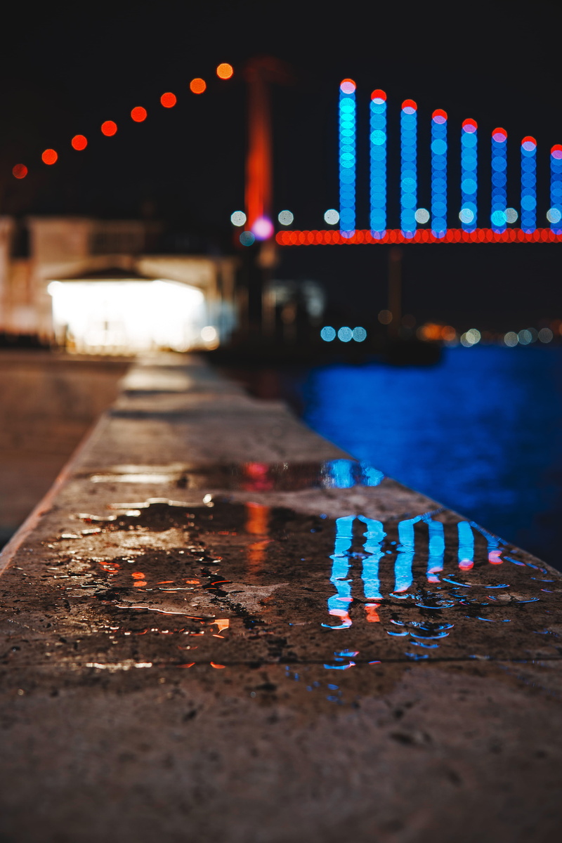 selective focus, surface level, dark, water, outdoors, building exterior, blur, transportation, pavement, lights, bridge, lighting equipment, lighted, evening