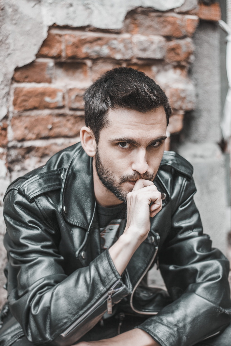 photo, portrait, front view, handsome, facial hair, posing, finger, sitting, young men, young adult, clothing, jacket, brick, architecture, biting, lifestyles