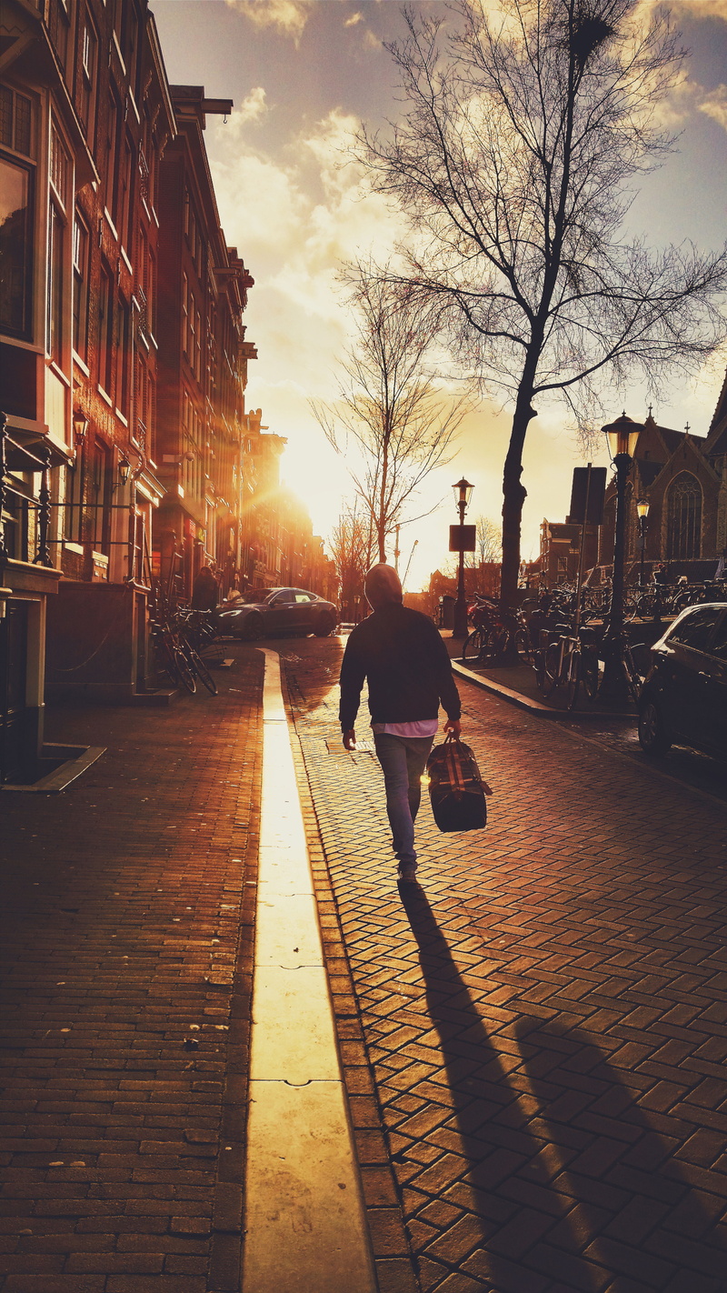 carrying, street, real people, sidewalk, golden, building exterior, roads, outdoors, trees, city street, one person, lens flare, nature, buildings, duffle bag