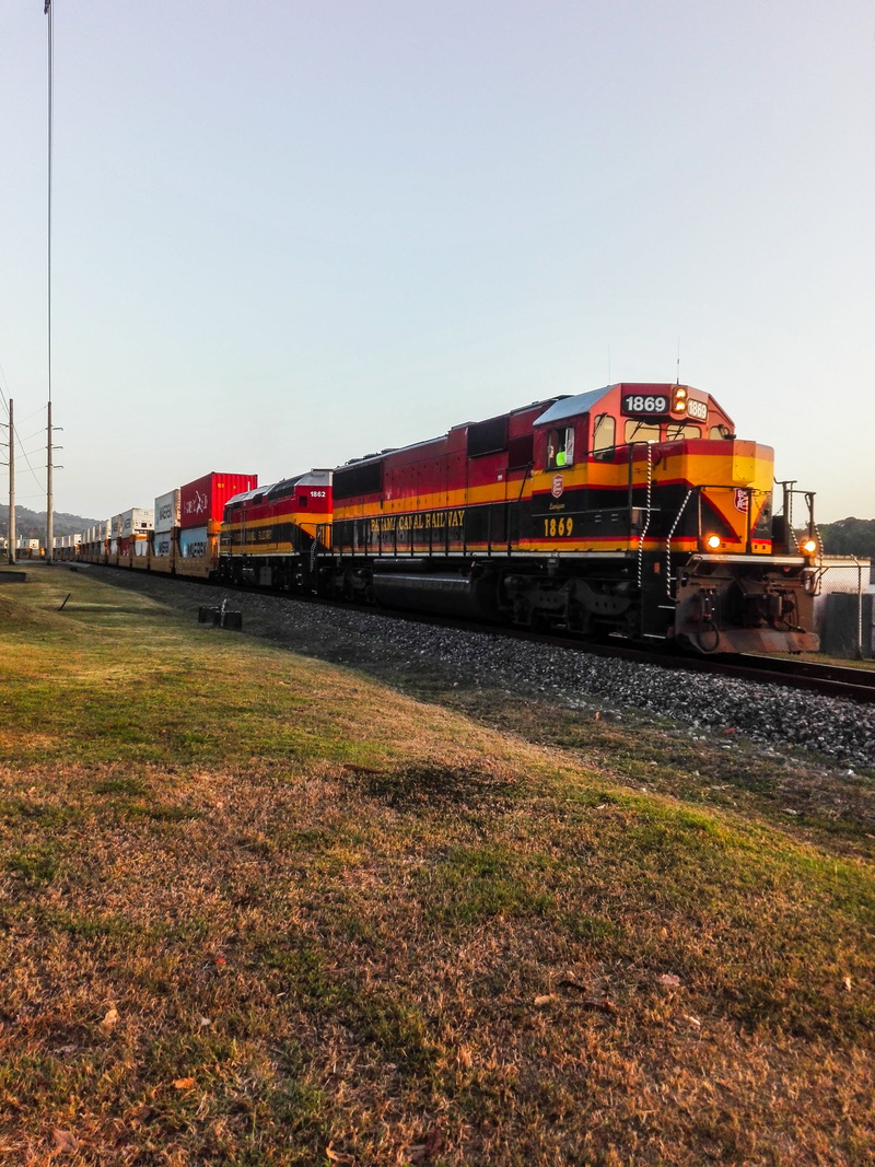 train track, shunting yard, outdoors, rail, locomotive, spoke, train, building, shipping container, orange, business, dirt road, sky, machine, railway