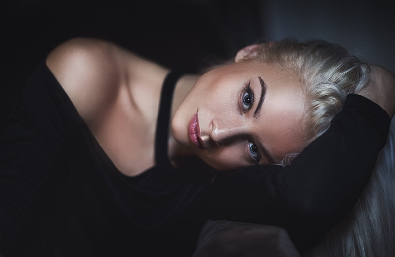 hair, bed, dark, blue eyes, face, black background, looking at camera, hairstyle, waist up, blonde, adult, 1080P, indoors, beauty, lying down, tanned