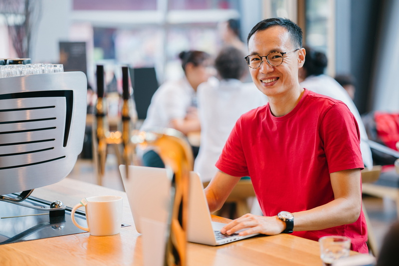 sitting, emotion, looking at camera, table, eyeglasses, men, using laptop, facial expression, portrait, adult, mug, casual clothing, blurred background