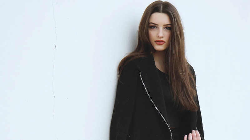 wall - building feature, coats, contemplation, looking at camera, front view, black clothing, women, brown hair, teenager, hairstyle, white wall, casual clothing
