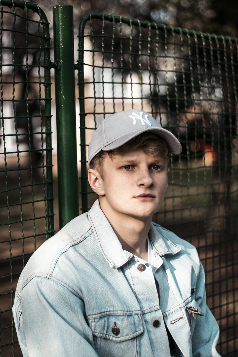 hat, school, day, beside, headshot, sitting, cap, face, architecture, jacket, portrait, security, chainlink fence, one person, looking at camera, sad, pants