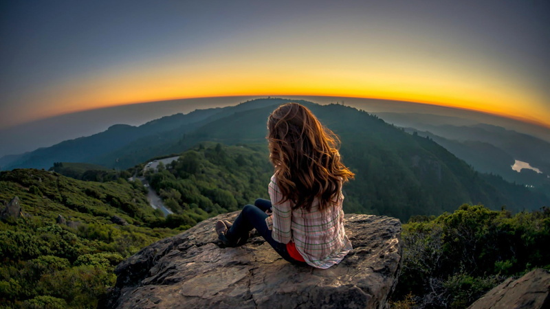 outdoors, 1080P, sleeved, model, rear view, mountain range, sitting, real people, s, hair, sky, shirt, white, looking at view, long hair, mountain, panorama