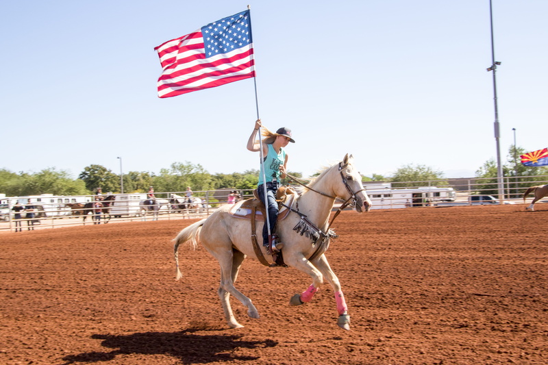 sign, women, shadow, tree, adult, rider, full length, rodeo, domestic animals, riding, sport, racer, carrying, holding, fence, girl on horse, day, patriotism