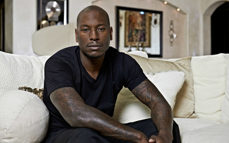 men, portrait, looking at camera, focus on foreground, sitting, real people, one person, living room, three quarter length, contemplation, tattoo, Tyrese Gibson