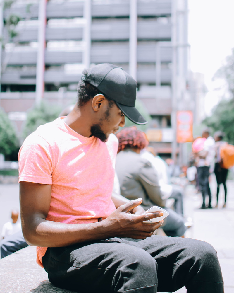 technology, holding, cap, sitting, incidental people, man, t-shirt, people, focus on foreground, bench, adult, phone, casual wear, urban, wear, three quarter length