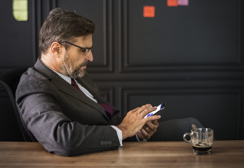 westerner, facial hair, indoors, checking, glass, man, business, technology, glasses, sitting, photo, smart phone, mature adult, 2K, mug, suit, black