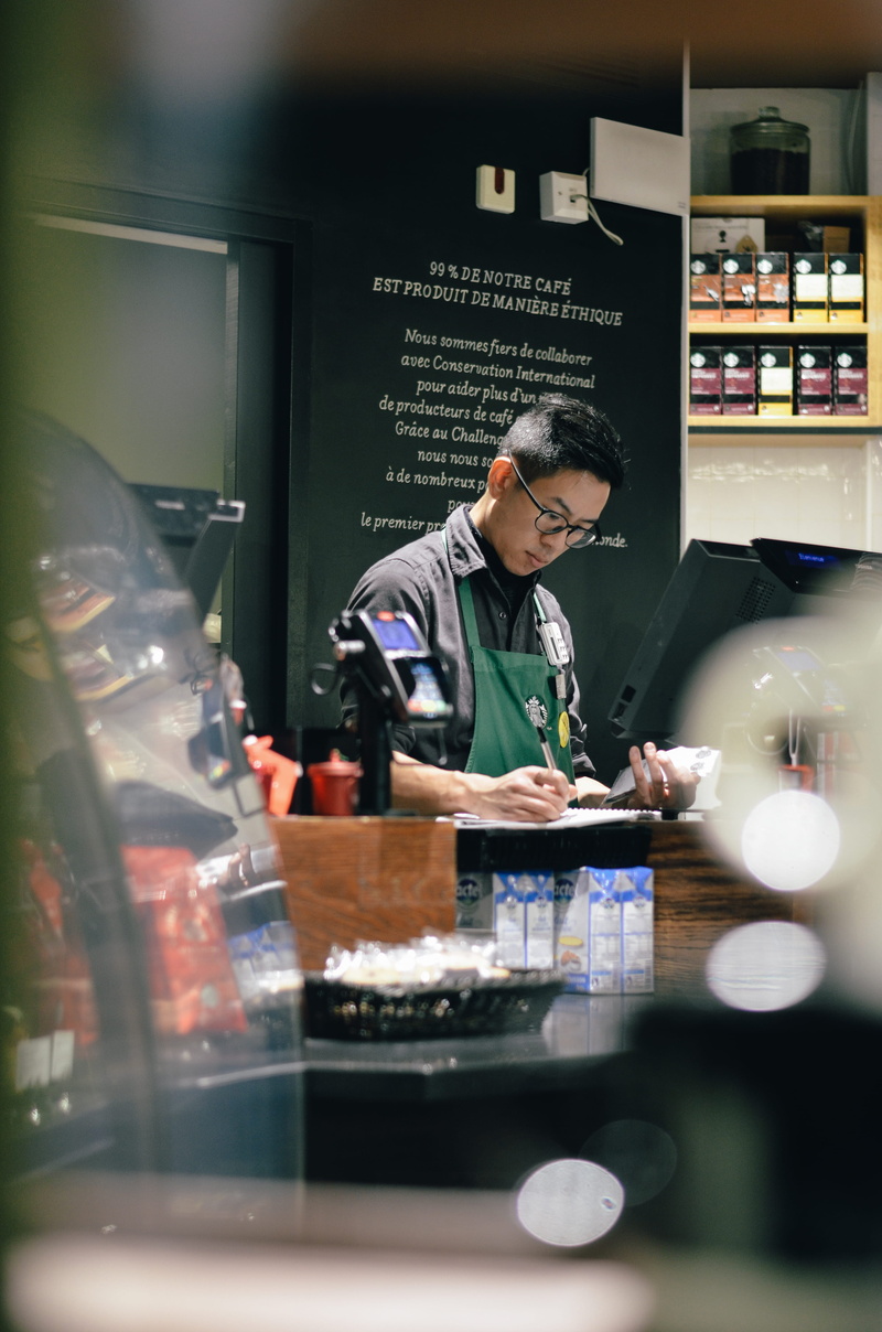person, shelf, adult, human, small business, waist up, one person, barista, bar counter, Starbucks Coffee, menu, machine, connection, store, standing, finger