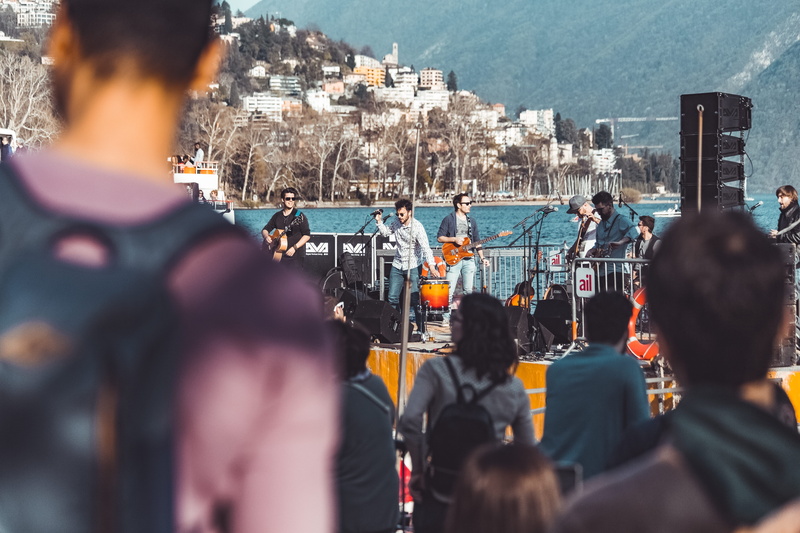 guitar, built structure, crowd, city, musicians, focus on background, song, architecture, nature, performance, men, singer, real people, standing, day