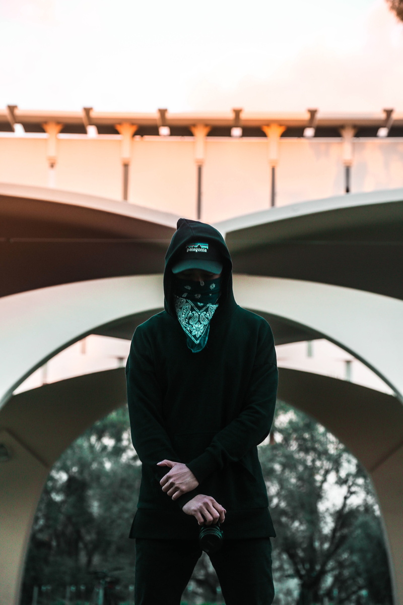 man, looking at camera, front view, outdoors, hoodie, mask - disguise, lifestyles, one person, symmetry, street, portrait, three quarter length, building