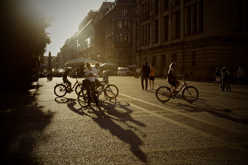city life, transportation, buildings, sunlight, person, silhouette, people, men, bicycles, land vehicle, city, pavement, shadow, architecture, bikes, sport
