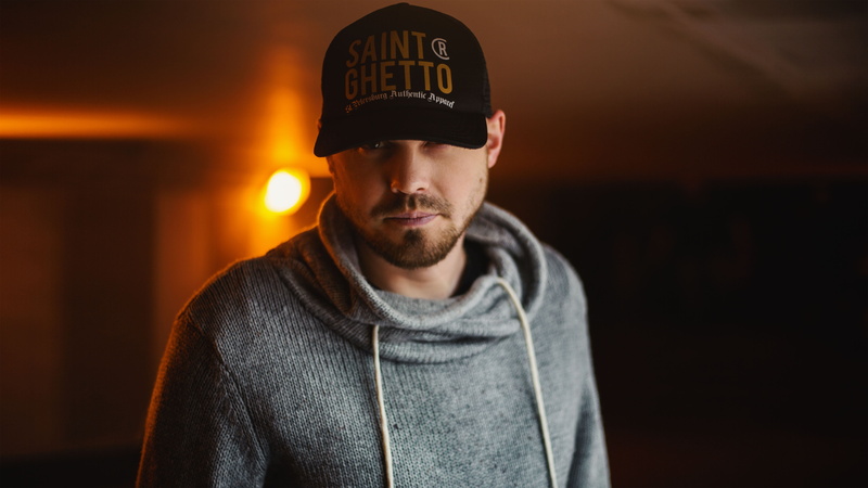 headshot, cap, looking at camera, waist up, jacket, hat, facial hair, beard, clothing, night, one person, light, casual clothing, wearing, hoody, architecture