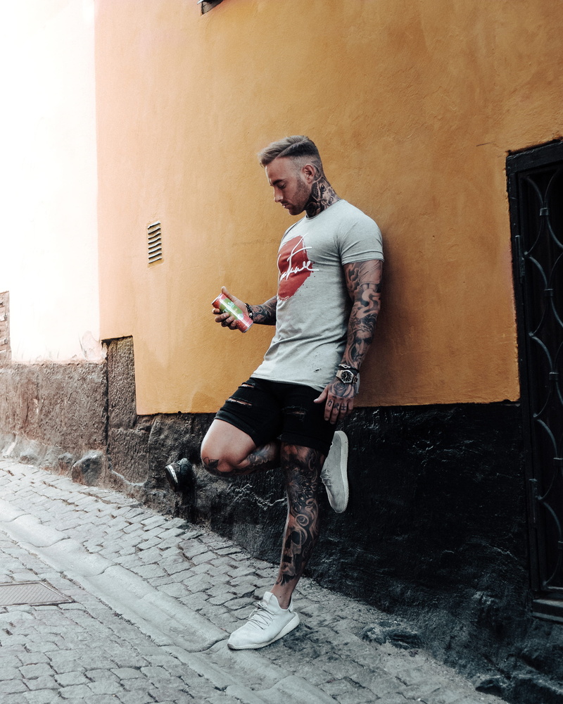 casual clothing, building exterior, built structure, leaning, leg tattoo, inked, street, portrait, looking down, man, wall, watch, leisure activity, full length