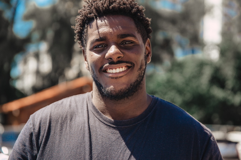 man, black, headshot, looking at camera, lifestyles, curly hair, daytime, happiness, tree, adult, black man, focus, wearing, emotion, hairstyle, young adult