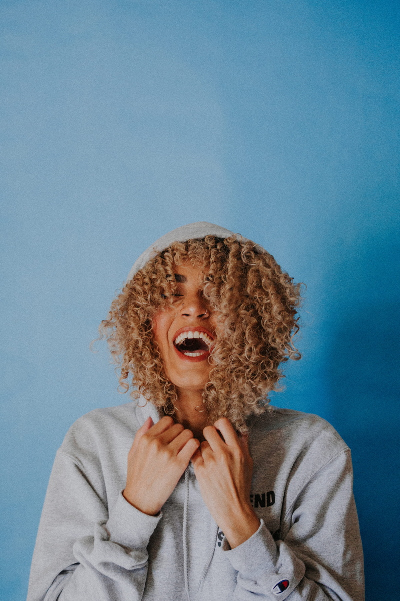 gray, laugh, blue, casual clothing, mouth open, smiling, sweater, woman, curly hair, wall - building feature, front view, portrait, hair, smile, wearing