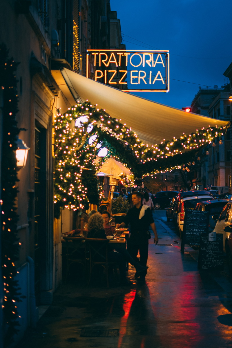 pavement, street, city, pizzeria, evening, commerce, lighting equipment, chairs, downtown, christmas lights, real people, walking, hanging lights, dark
