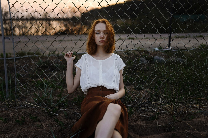 looking at viewer, women outdoors, day, redhead, one person, blouses, 2K, sitting, hairstyle, three quarter length, outdoors, women, contemplation, chainlink fence