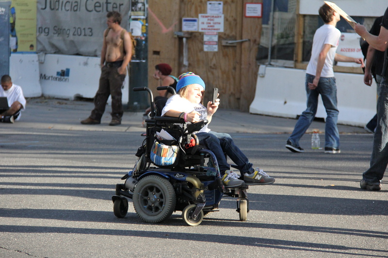 wheel, asphalt, men, vehicle, incidental people, sign, human, wheelchair, motorized, chair, furniture, pants, person, denim, road marking, footwear, architecture