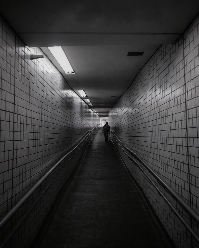 subway system, 2K, one person, dark, walking, underpass, underground walkway, building, flooring, lighting equipment, illuminated, black and white, diminishing perspective