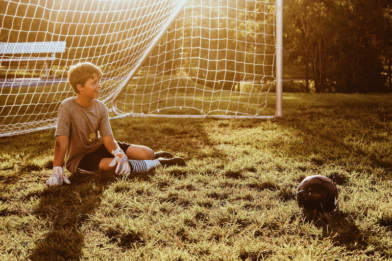boy, human, grass, person, team, people, sphere, young adult, sitting, sun, one person, ball, plant, team sport, full length, casual clothing, kicking