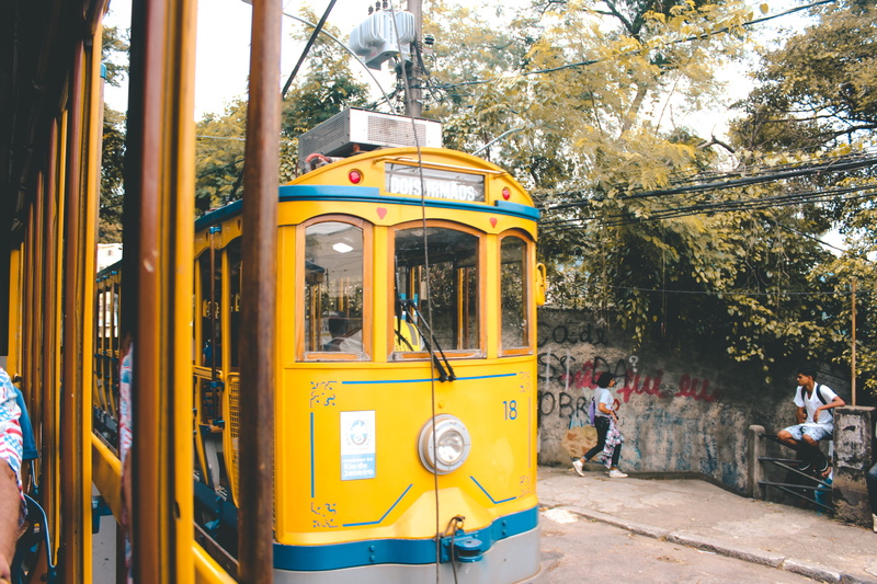 people, incidental people, wall, tram, standing, land vehicle, outdoors, beside, vehicle, group of people, mode of transportation, streetcar, person, flagstone