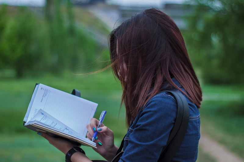 depth of field, adult, woman, pen, one person, waist up, close-up, brown hair, notebook, outdoors, holding, lifestyles, writing, casual clothing, hair