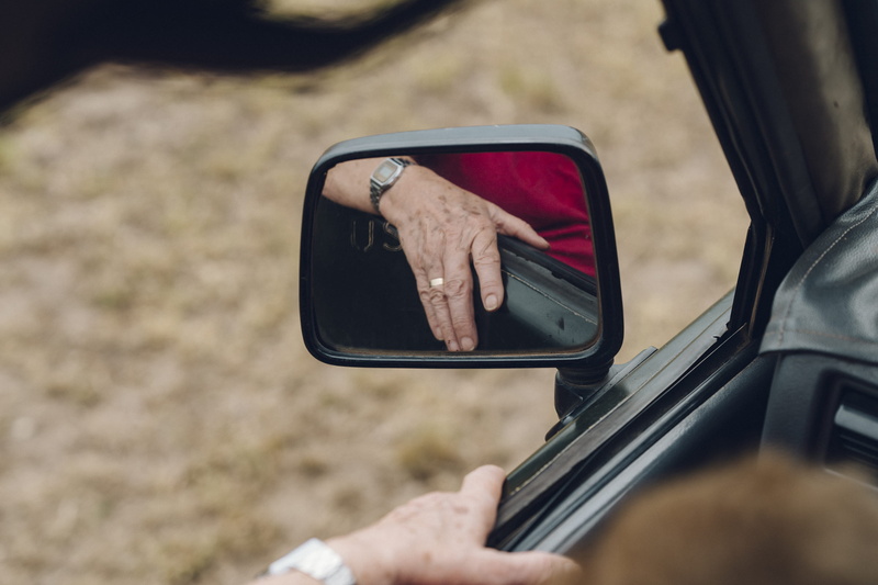 human, outdoors, building, selective focus, car, side-view mirror, boardwalk, day, cowbell, cell phone, technology, mode of transportation, hand, reflection