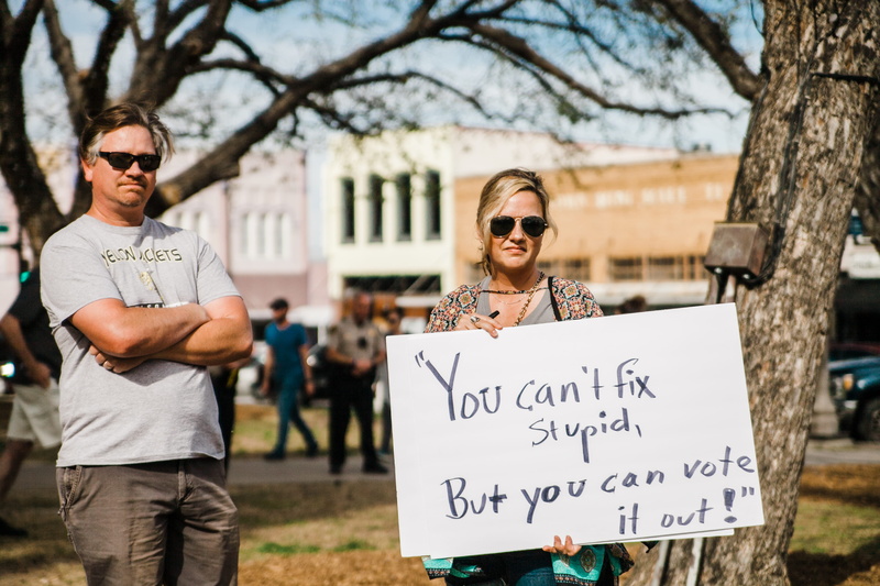 communication, nature, sign, board, holding, signage, buy, print, man, adult, can, standing, portrait, wearing, stupid, woman, text, white, tree, focus on foreground